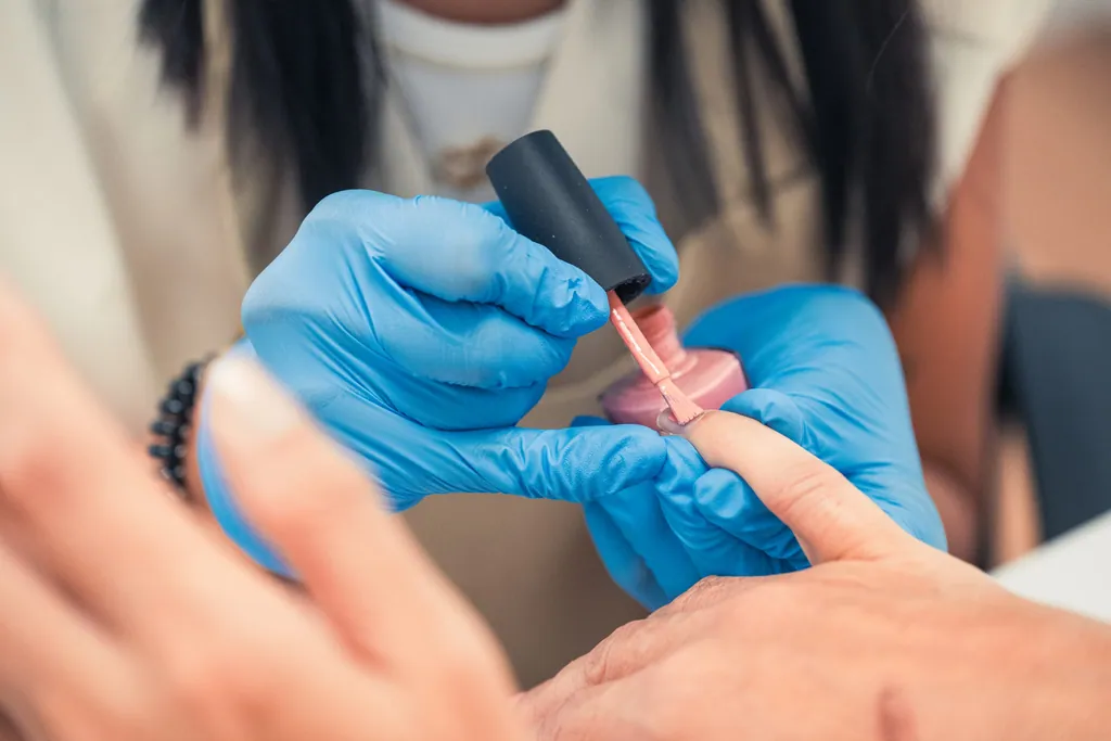 Profissional de luvas azuis a aplicar verniz cor-de-rosa nas unhas de uma cliente